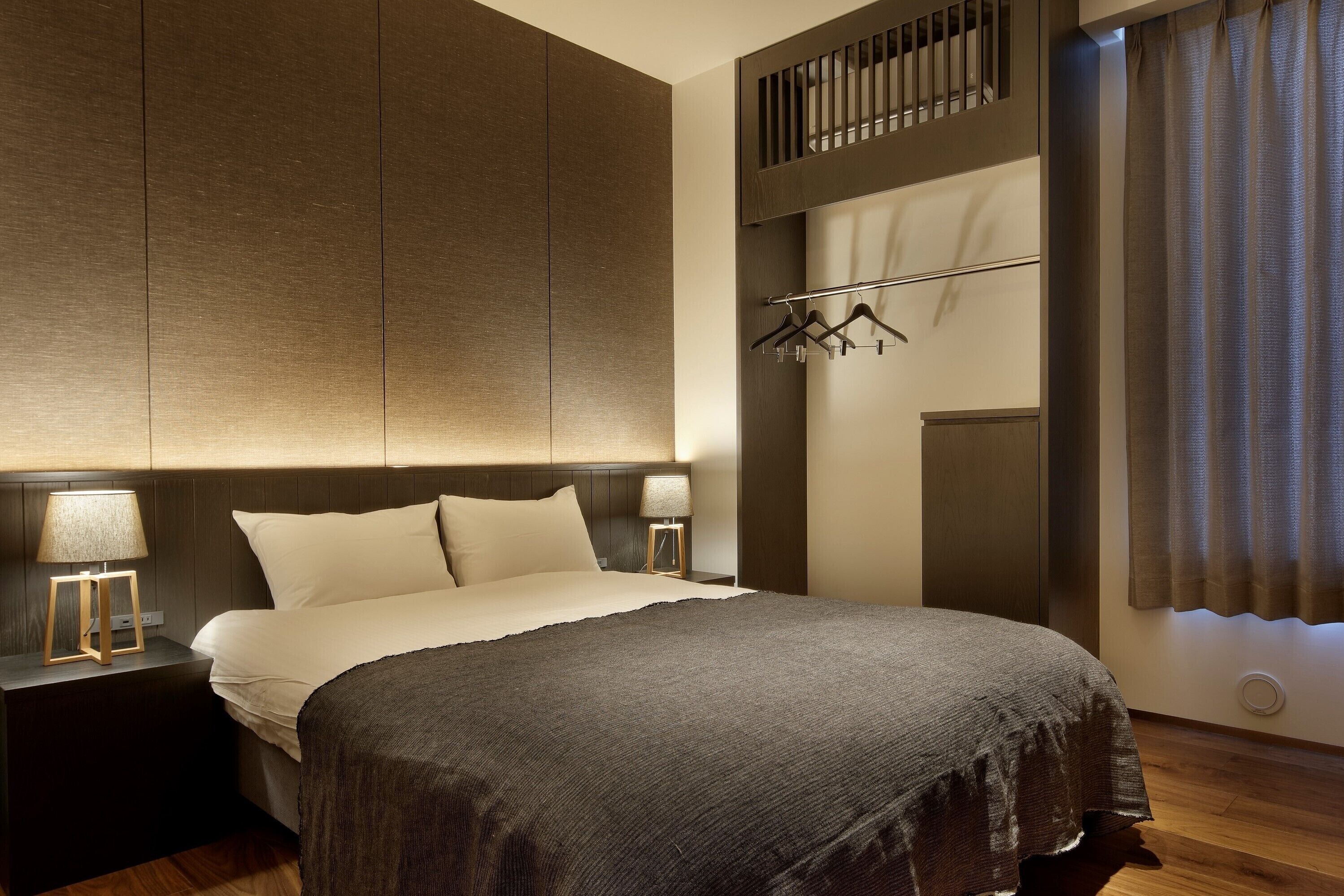 A modern hotel room with a large bed, a nightstand with a lamp, a wardrobe, and a coat rack.