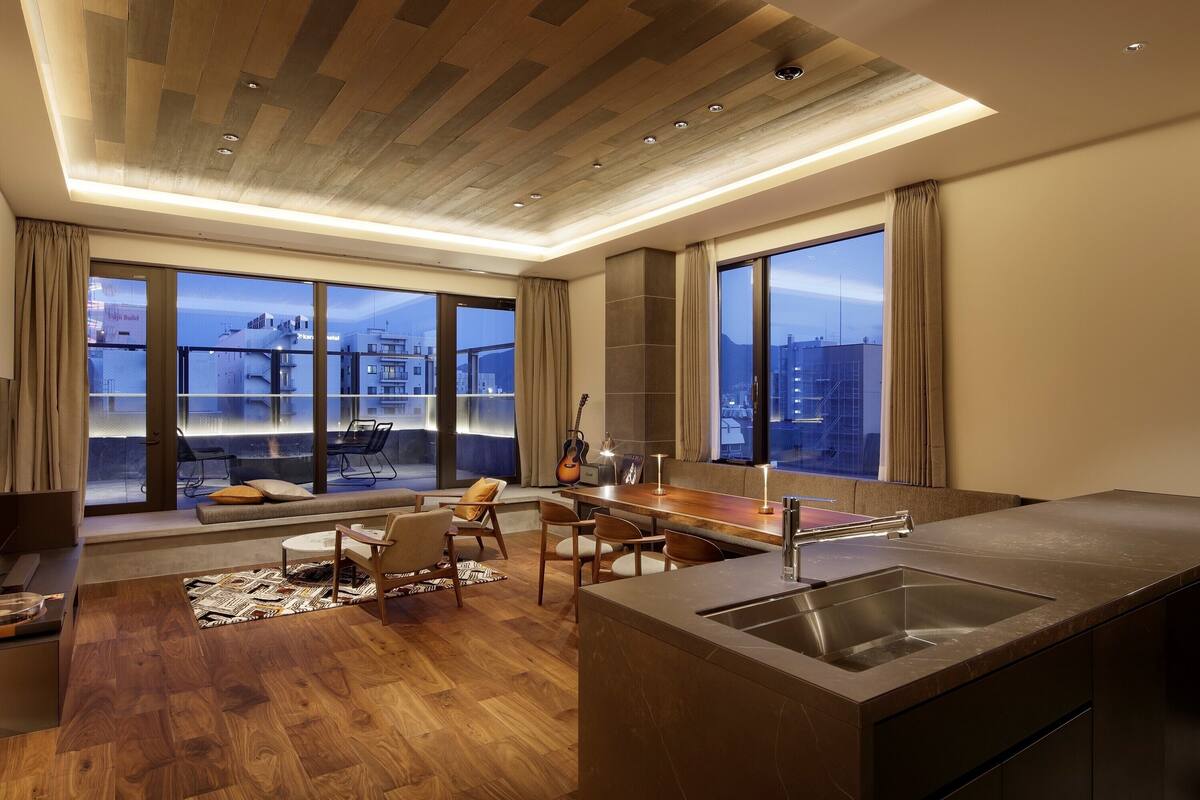 A modern living room with a wooden floor, a sofa, a dining area, and a kitchen with a large window.