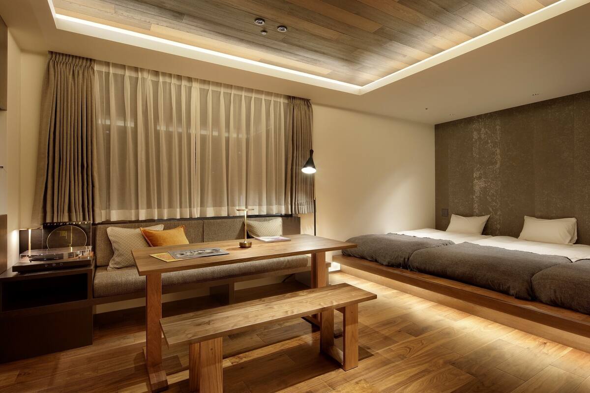 A modern room with a wooden floor, a sofa bed, a wooden table with a bench, a desk with a lamp, and a large window with curtains.
