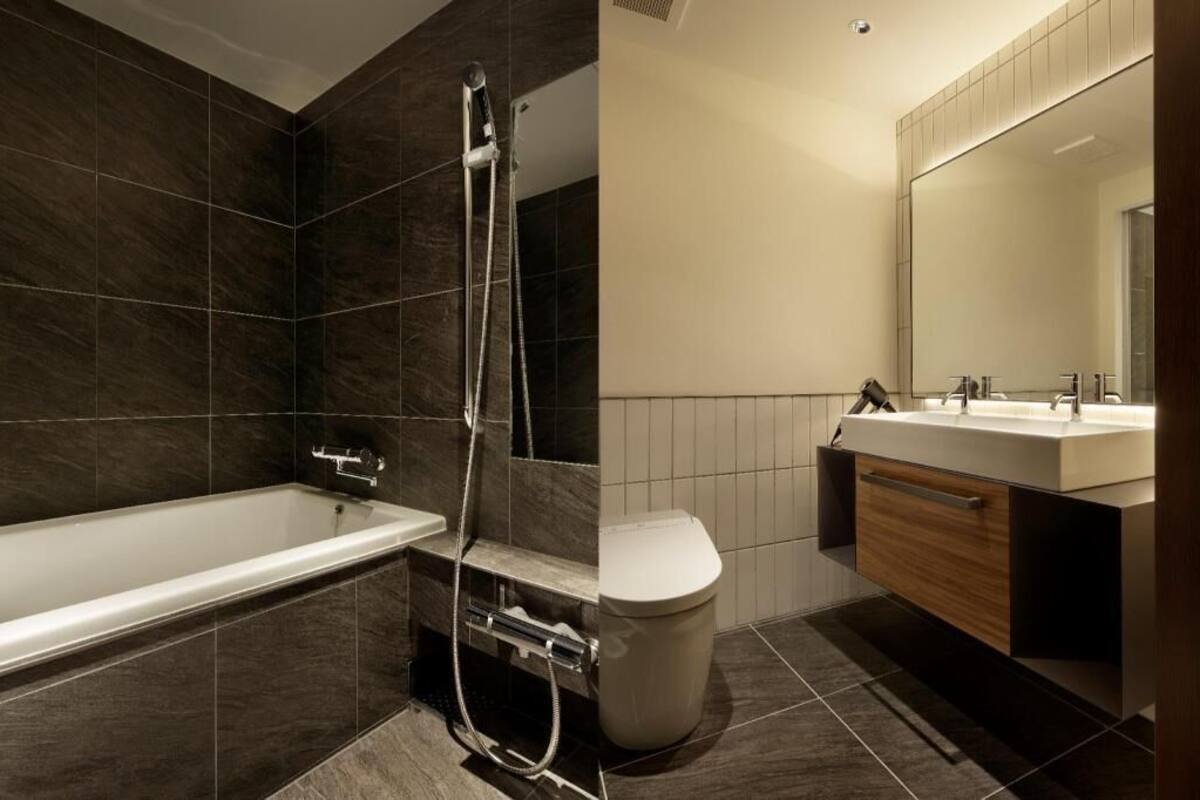 A modern bathroom with a bathtub, shower, and toilet, featuring dark tiles and a wooden vanity.