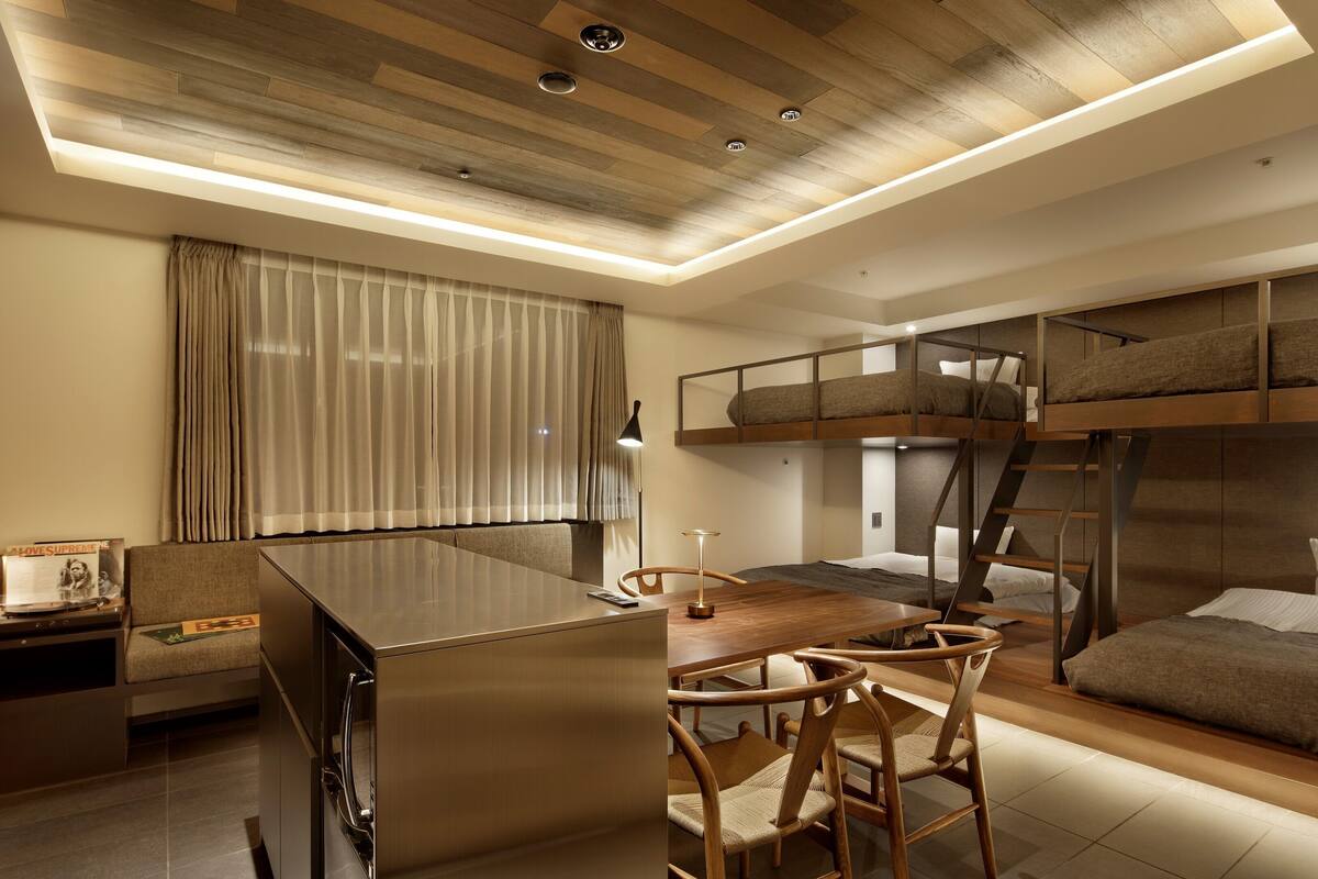 A compact hotel room with a bunk bed, a kitchenette, a dining area, and a large window with curtains.