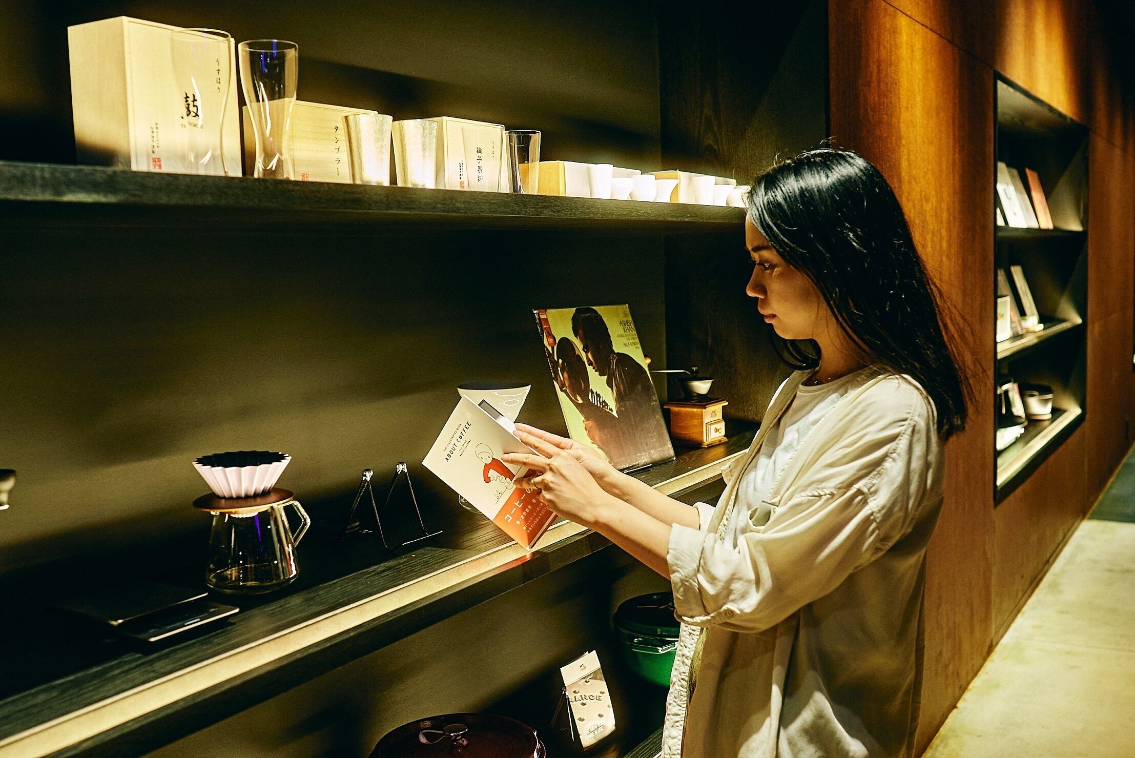Eine Person liest ein Buch in einem Raum mit Bücherregalen, einer Kaffeemaschine und einem gerahmten Bild.