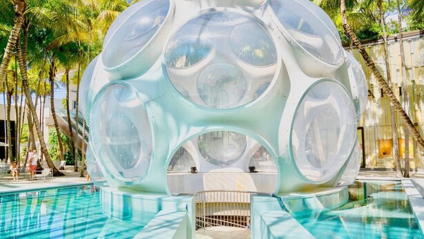 Une structure sphérique transparente, dotée de plusieurs ouvertures circulaires, située dans un espace piscine entouré de palmiers.