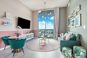 A modern living room with a large window, a flat-screen TV, teal chairs, a green sofa, and a small round table.