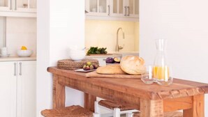 A kitchen with white cabinets, a wooden table with bread, fruit, and a glass bottle. - The view 2 (Santa Maria Paros Νάουσα)