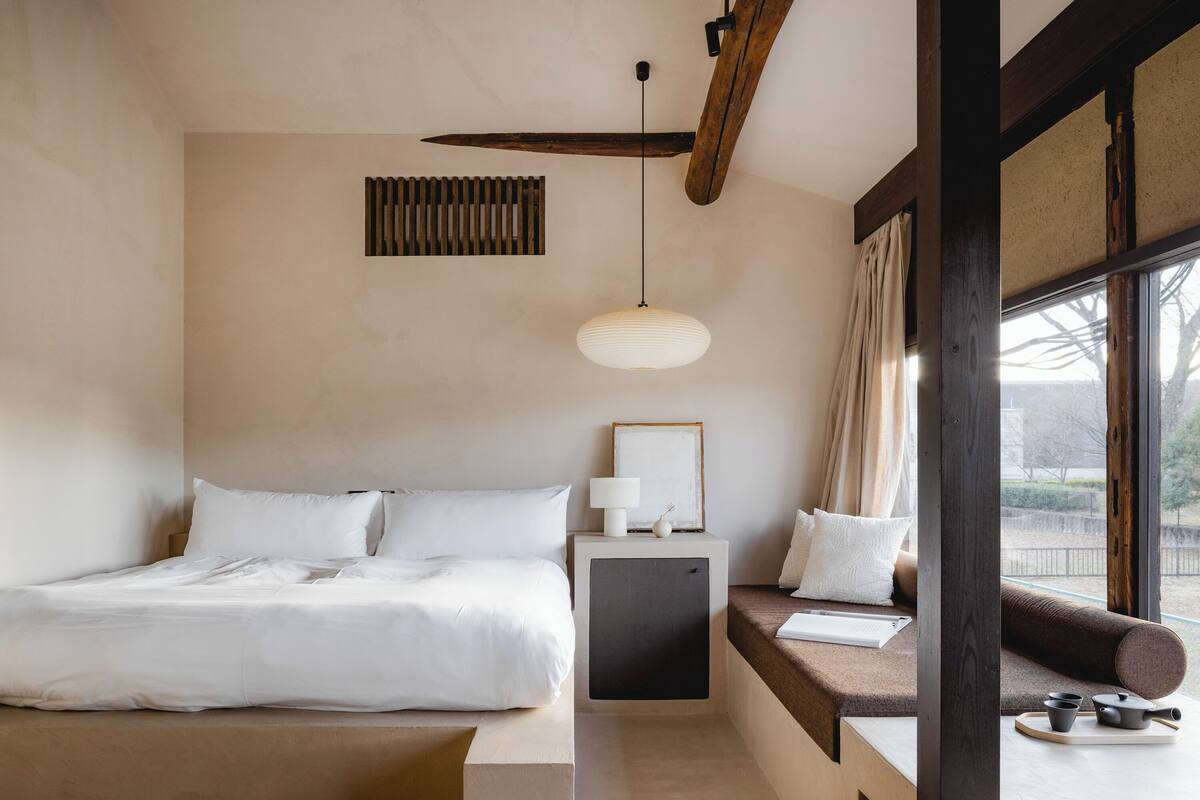 Ein schlicht eingerichtetes Hotelzimmer mit Bett, Nachttisch, Lampe und Blick durch ein Fenster.