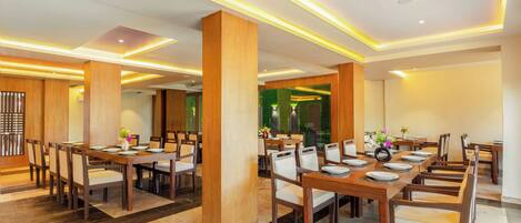 A spacious dining area with long wooden tables and chairs, featuring a modern ceiling design with recessed lighting.