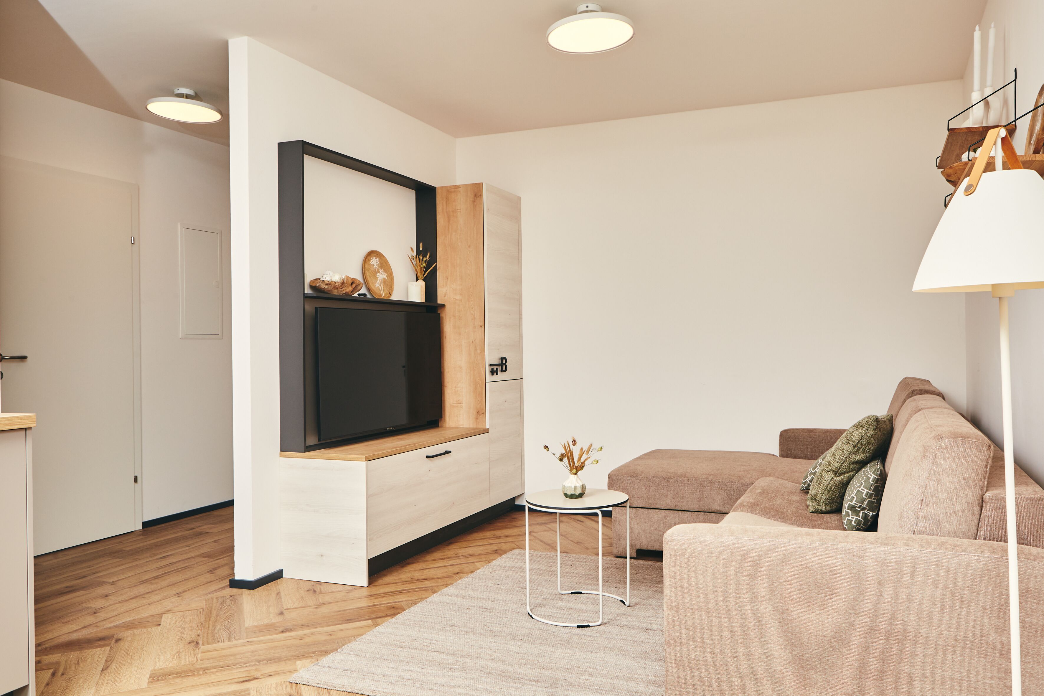 Una sala de estar moderna con suelo de madera, un sofá color beige, una pequeña mesita auxiliar blanca y un televisor de pantalla plana montado en un mueble de madera.