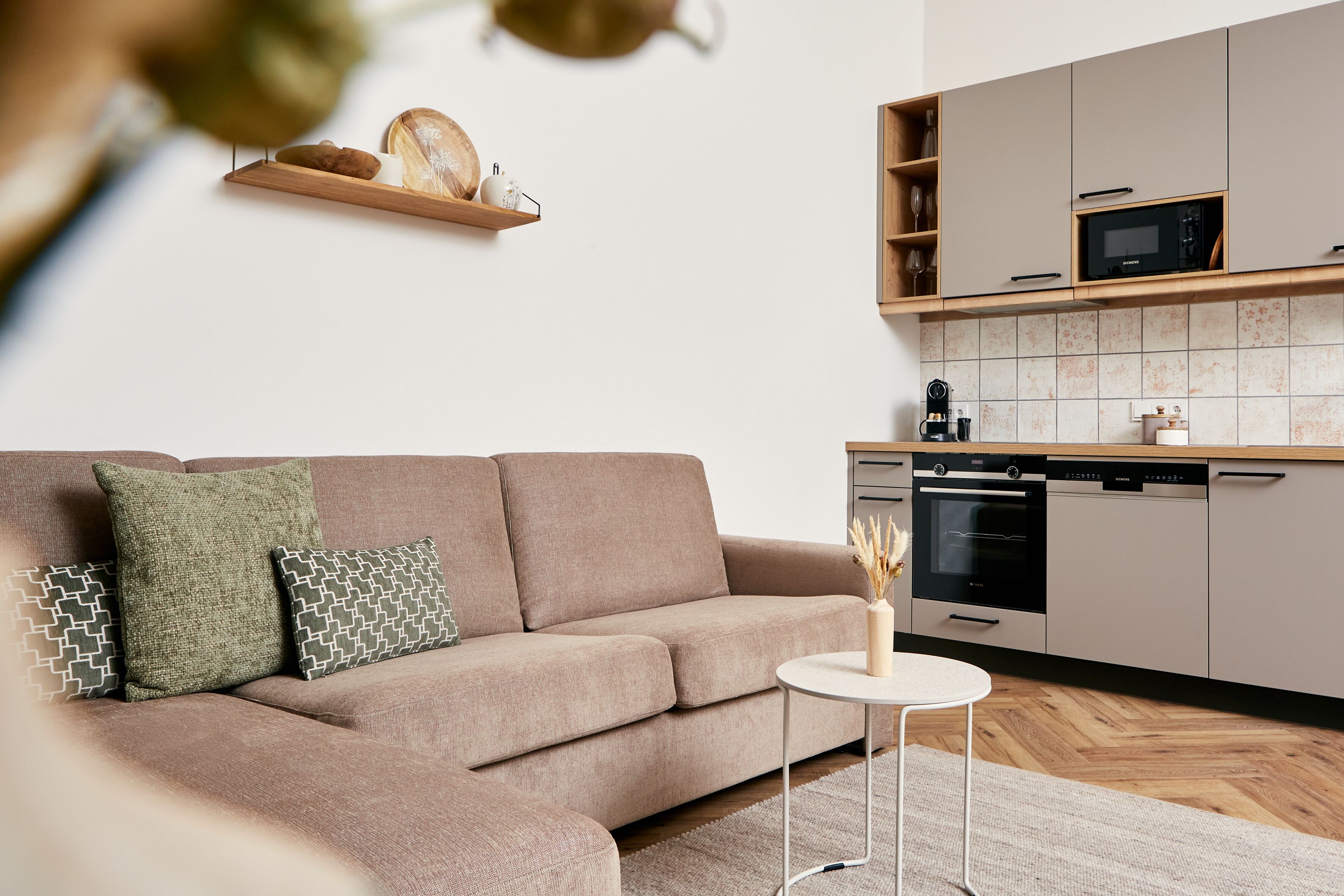 A modern living room with a beige sofa, a small round table, and a kitchen area with cabinets and appliances.