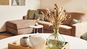 Ein Wohnzimmer mit einem braunen Sofa, einem kleinen runden Tisch, einer weißen Ablage mit Teekanne und einer Vase mit Trockenblumen.