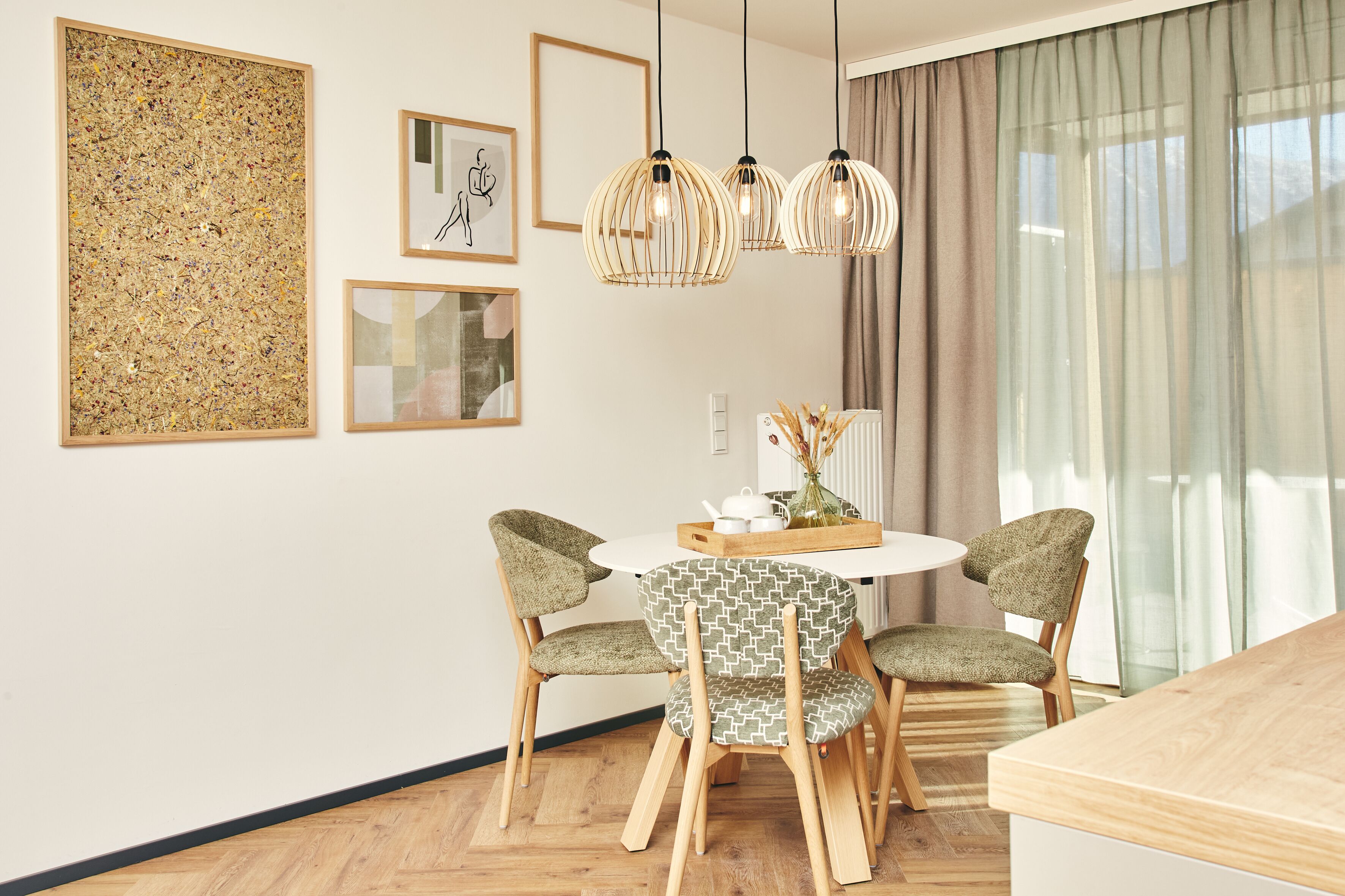Ein Essbereich mit einem runden Tisch, vier Stühlen und hängenden Pendelleuchten. An der Wand befinden sich gerahmte Kunstwerke und ein Fenster mit Vorhängen.