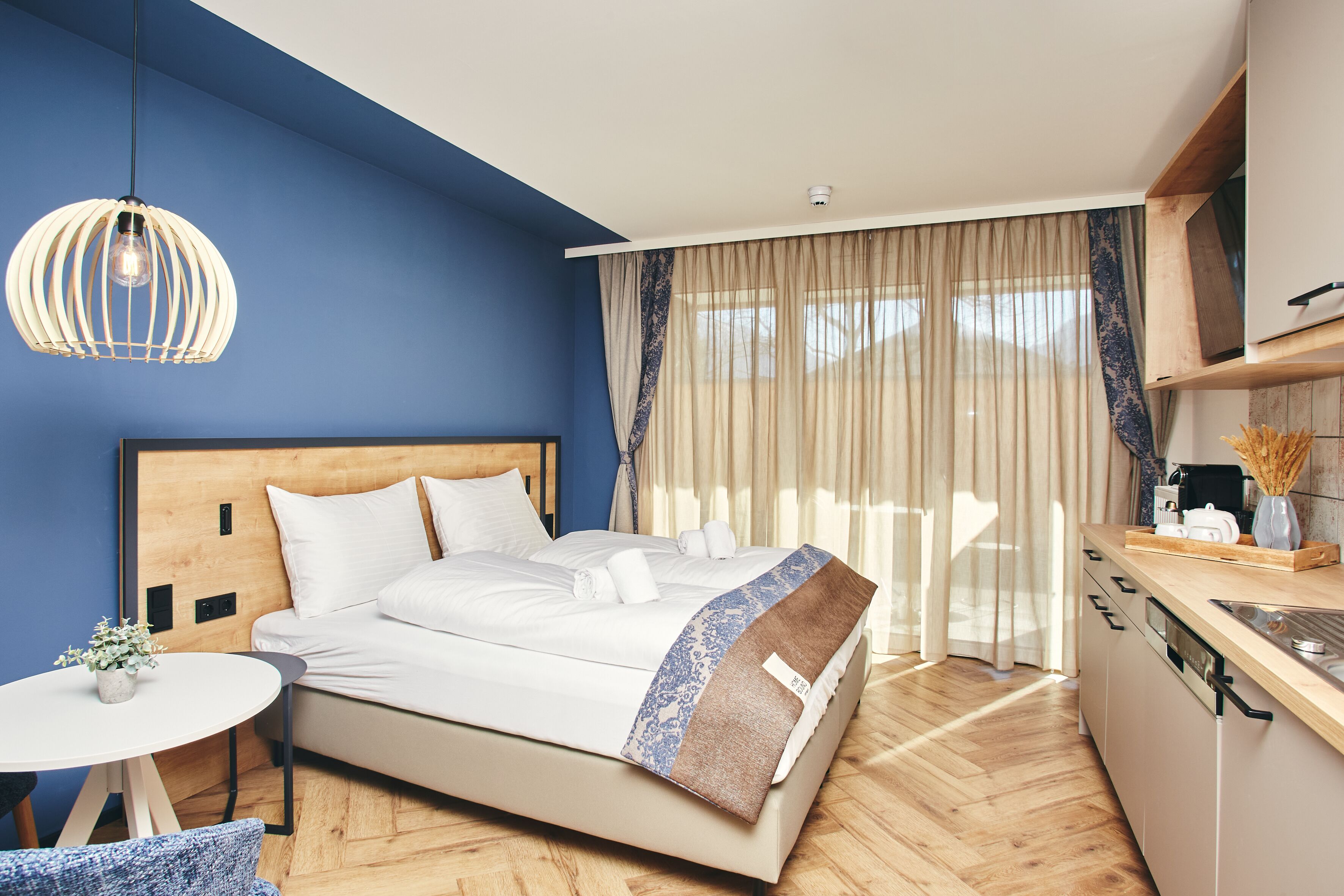 A modern bedroom with a wooden headboard, a white bed, a round table with a plant, and a kitchen area with a wooden countertop and cabinets.