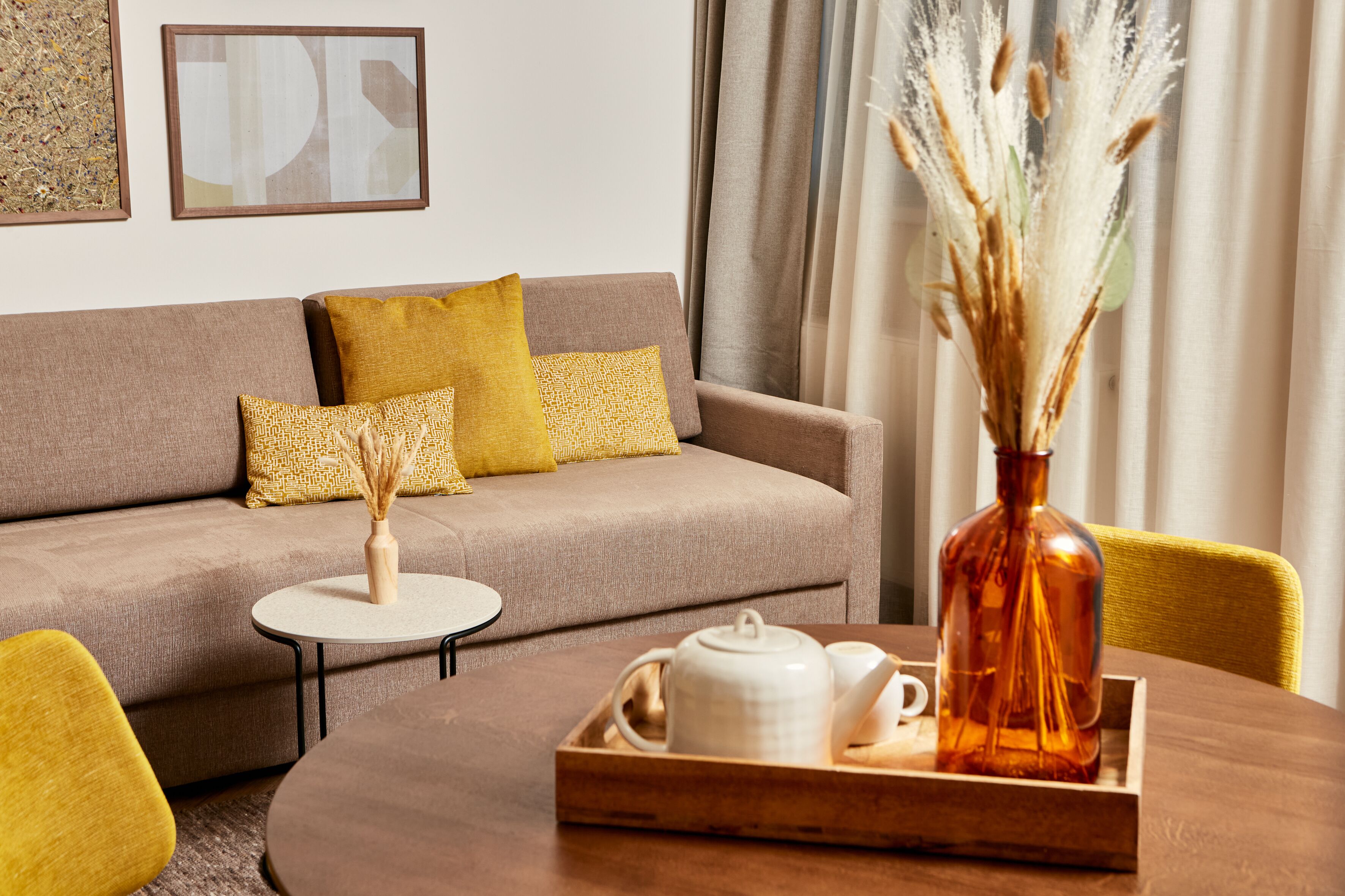 A modern living room with a sofa, yellow cushions, a round coffee table with a tray of tea set, and a vase with dried plants.