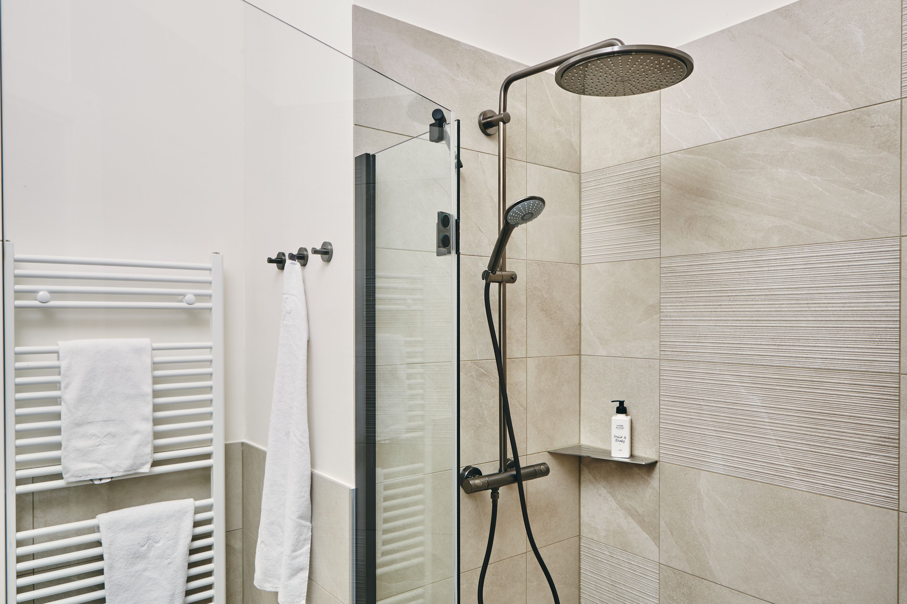A modern shower with a handheld showerhead and a fixed rain showerhead, a glass-enclosed shower stall, and a white towel rack with towels.