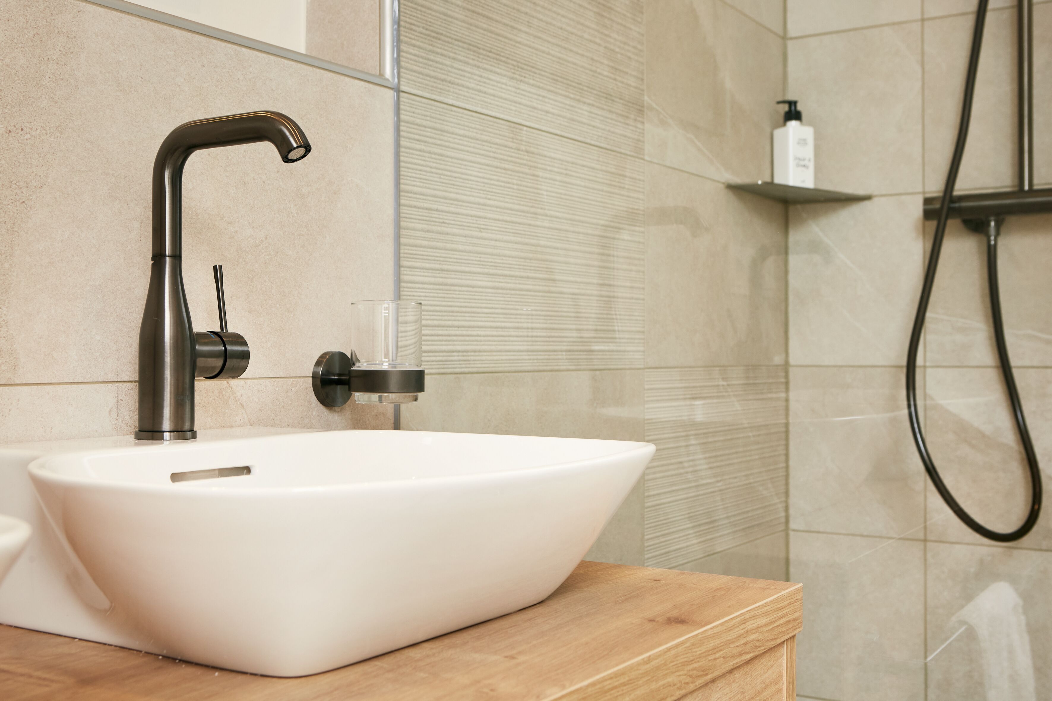 A bathroom with a white vessel sink, a modern faucet, a glass soap dispenser, and a shower area.
