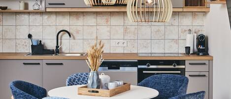 A modern kitchen with a round dining table, blue patterned chairs, wooden flooring, and pendant lights.