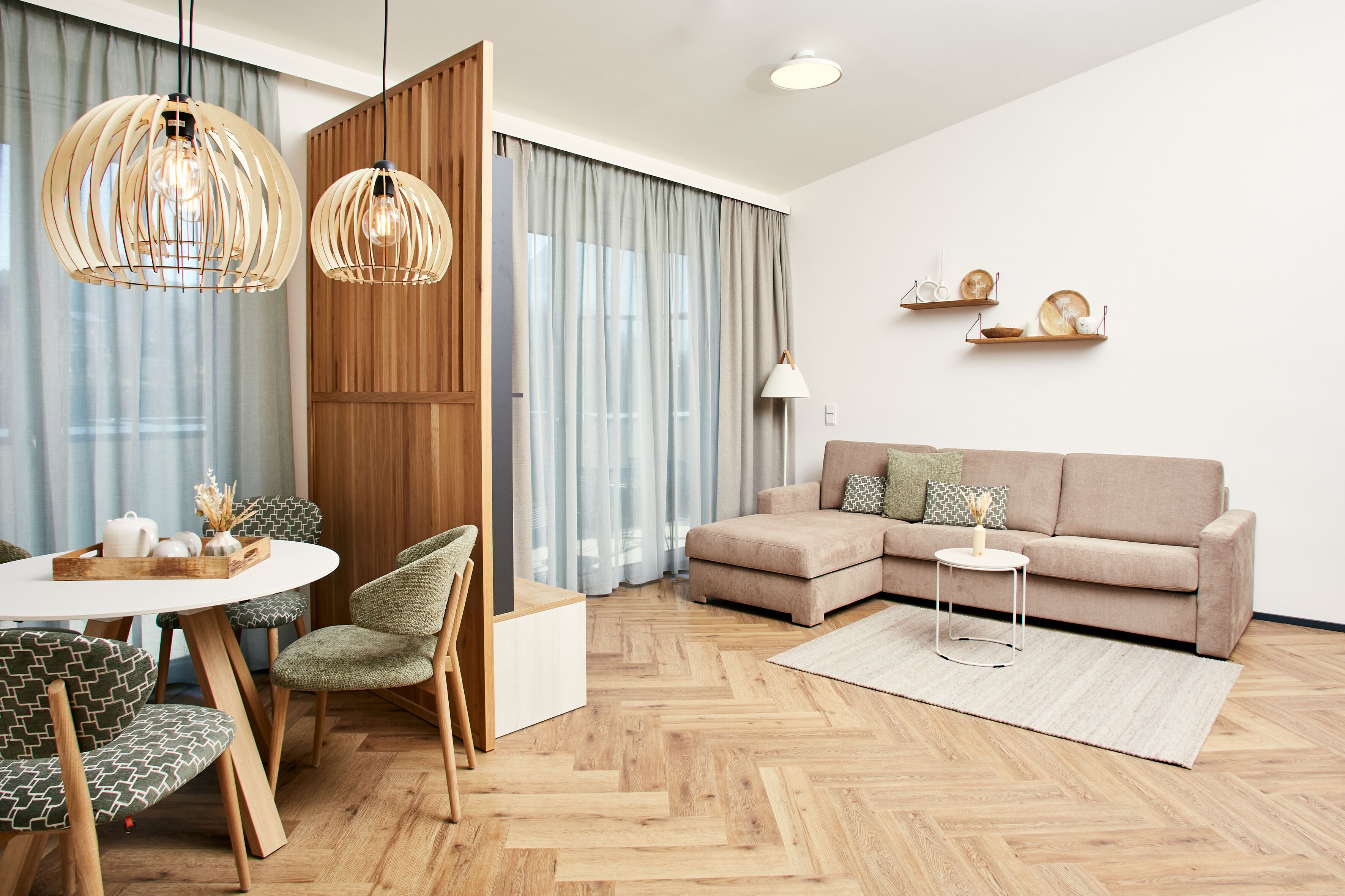 Una sala de estar moderna con piso de madera en patrón de espiga, un sofá seccional color beige, una mesa redonda con sillas y dos lámparas colgantes.