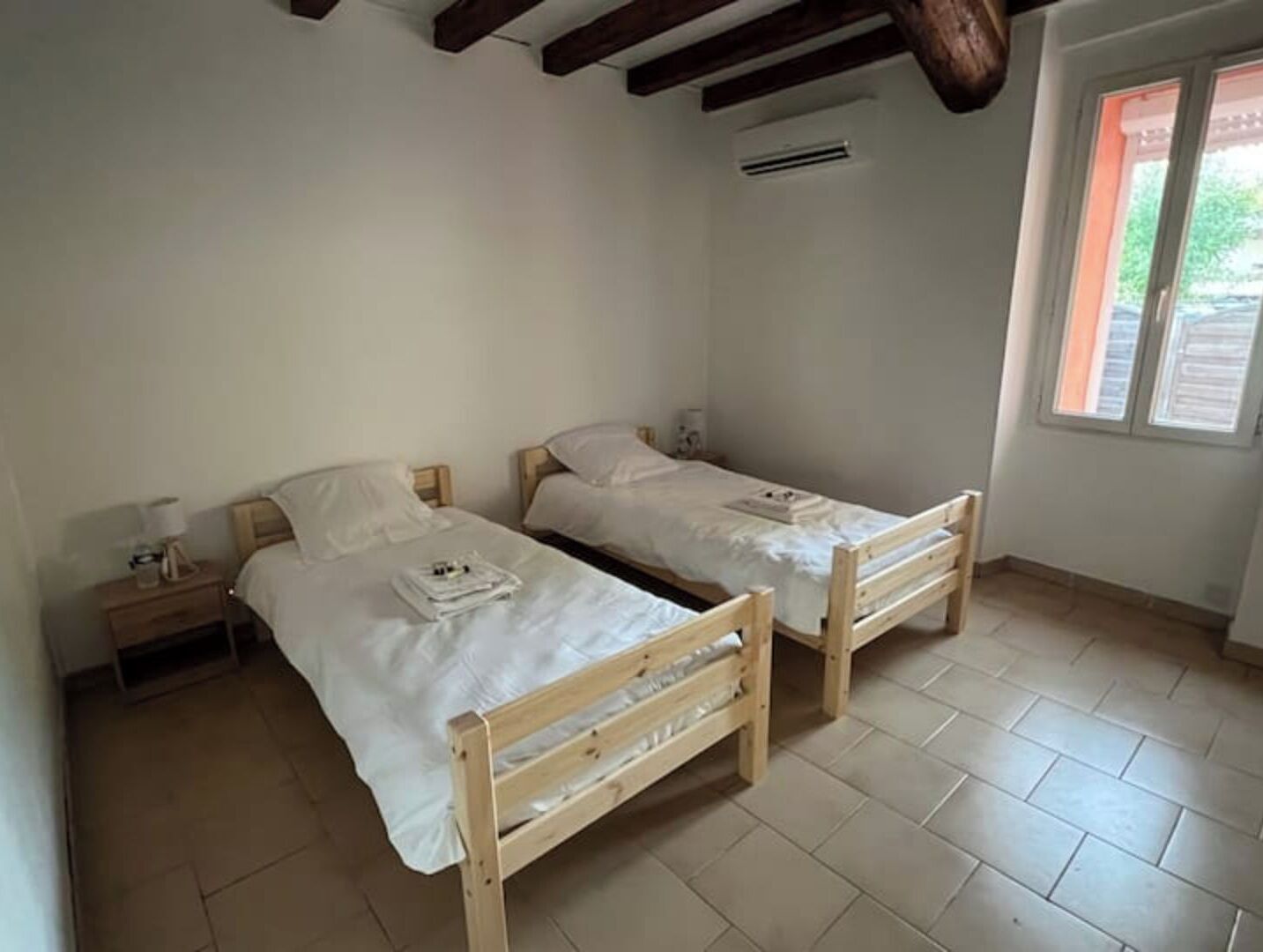 A small, clean room with two wooden beds, a nightstand, and a window with blinds.