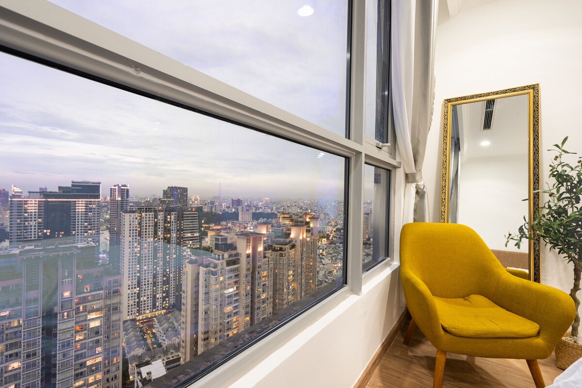 Quarto com uma janela grande que oferece vista para a cidade, uma poltrona amarela e um espelho na parede.