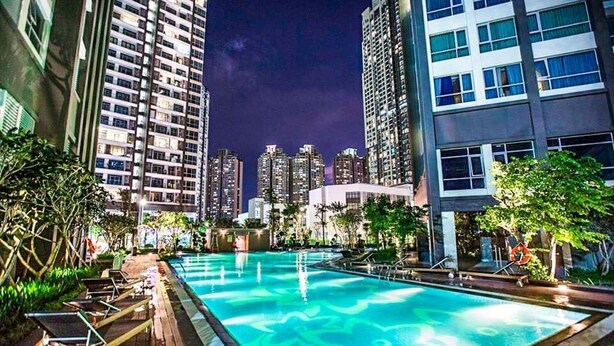 A nighttime view of a swimming pool area with lounge chairs, surrounded by tall buildings and greenery.