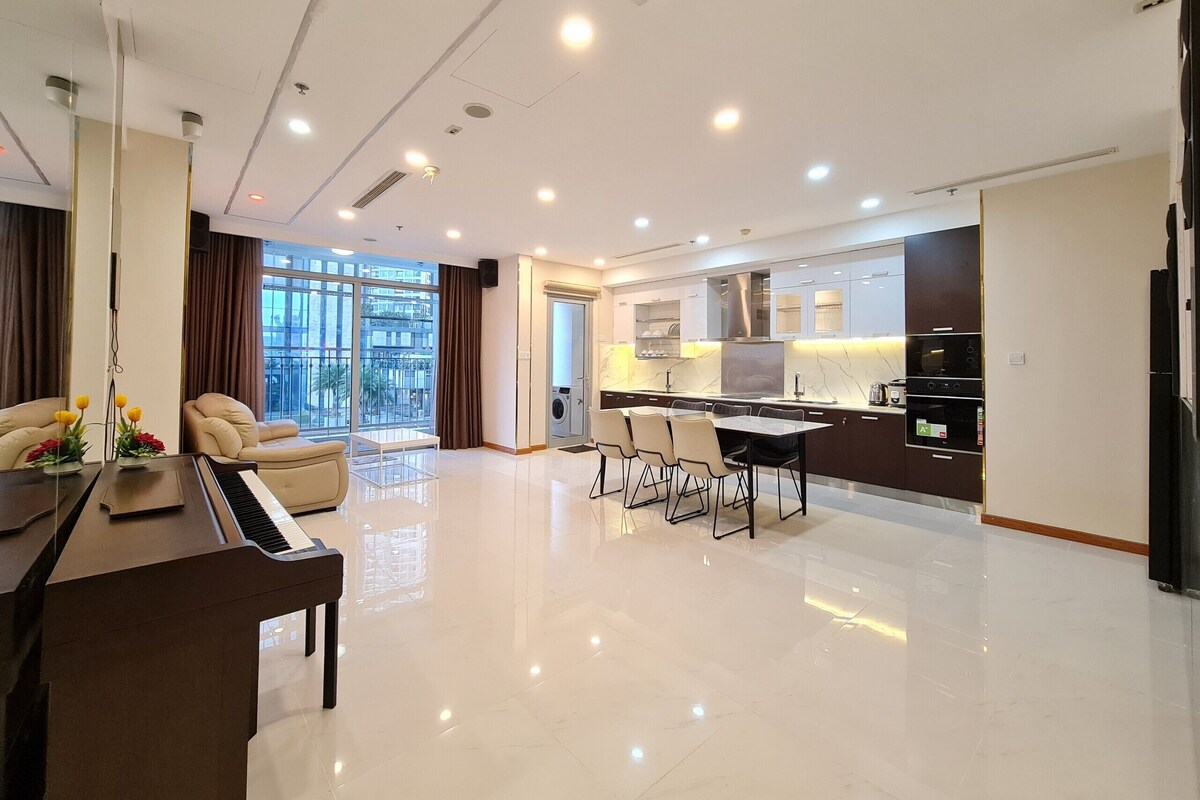 Uma sala de estar moderna com um piano, uma área de refeições e uma cozinha com armários escuros e bancada de mármore.