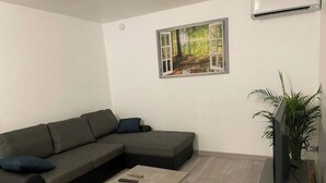 Ett modernt vardagsrum med en hörnsoffa, ett soffbord, en platt-TV och ett stort fönster med utsikt över en skog.