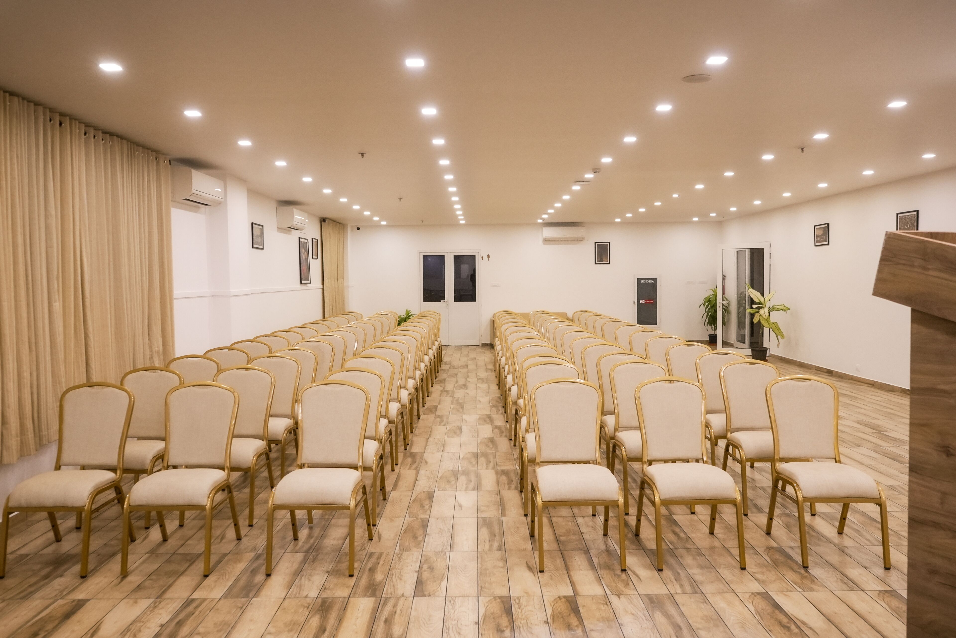 A conference room with rows of beige chairs and gold frames, wooden flooring, and recessed ceiling lights.