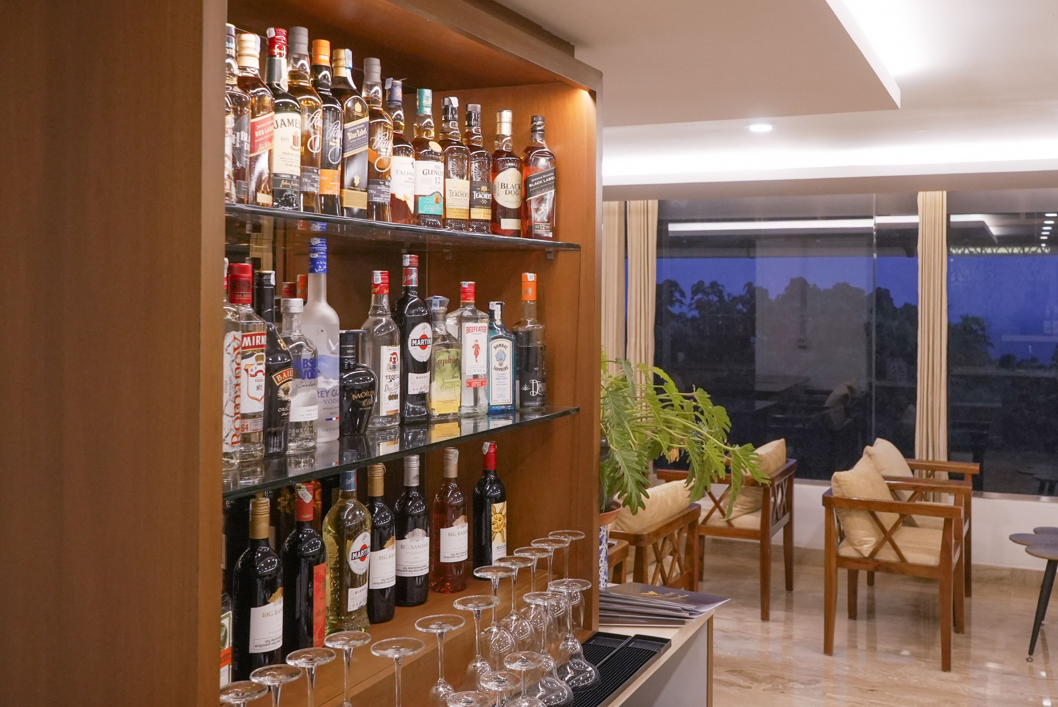 A bar area with shelves of liquor bottles and glasses, wooden chairs, and a view through a large window.