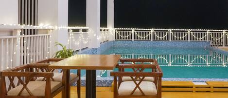 Una terraza de madera con piscina, barandillas blancas y luces colgantes.