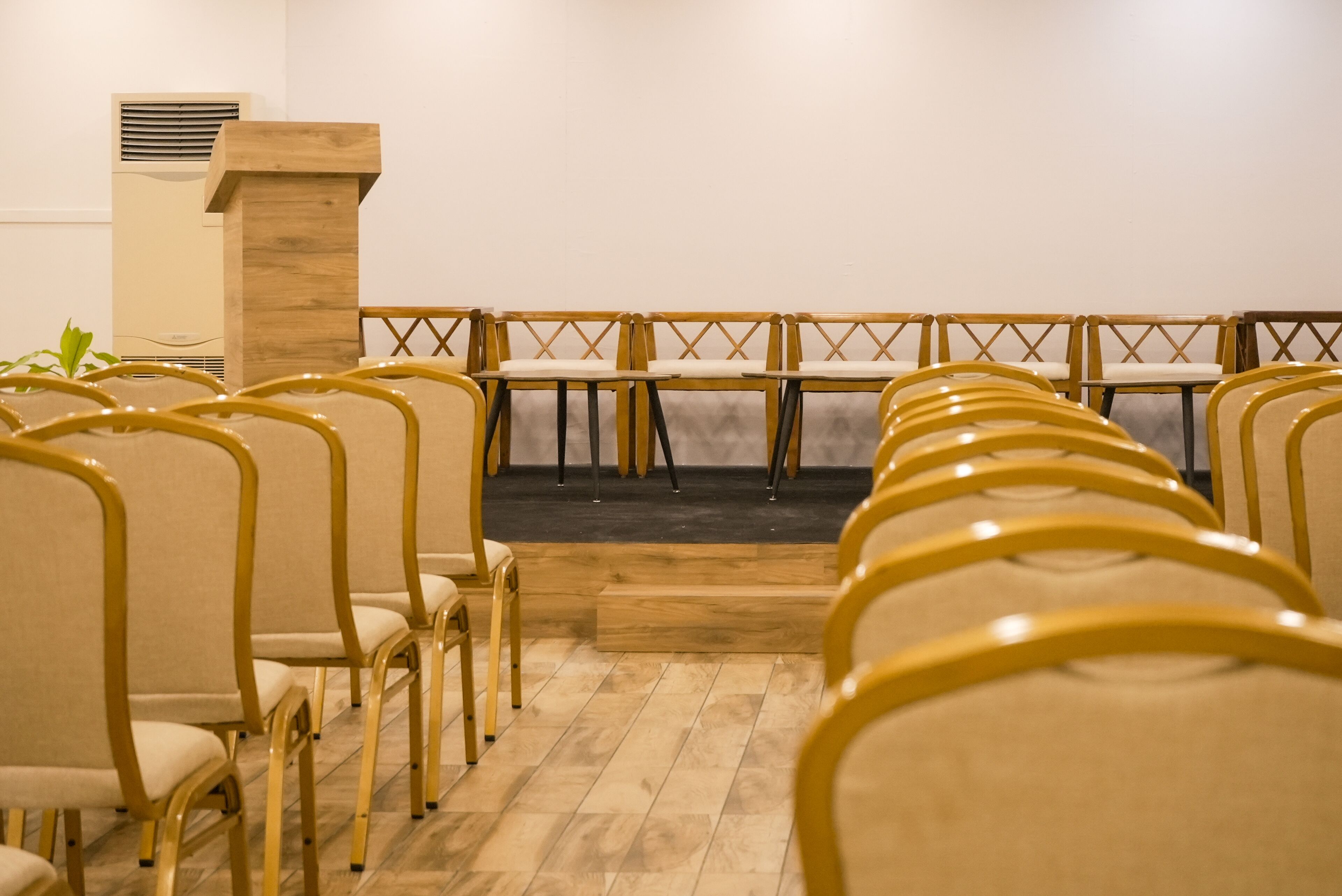 A row of yellow chairs with wooden frames and beige cushions, arranged in a semi-circular pattern facing a stage with a wooden podium.