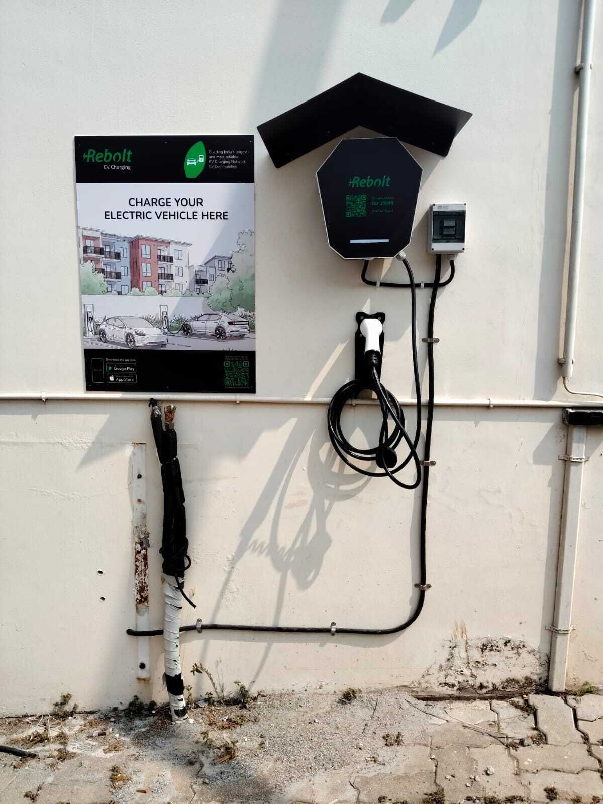 An electric vehicle charging station with a Rebolt sign, a charging cable, and a QR code for app download.