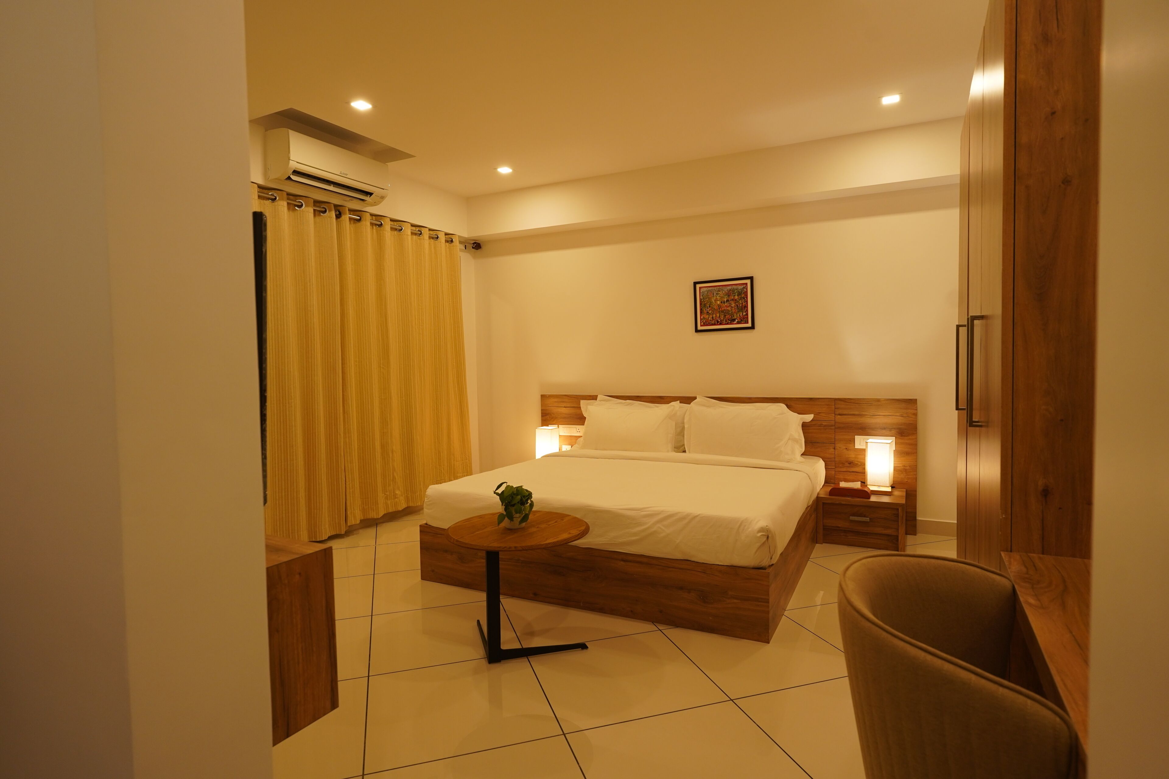 A modern hotel room with a wooden bed, a small round table, a chair, and a nightstand with a lamp.