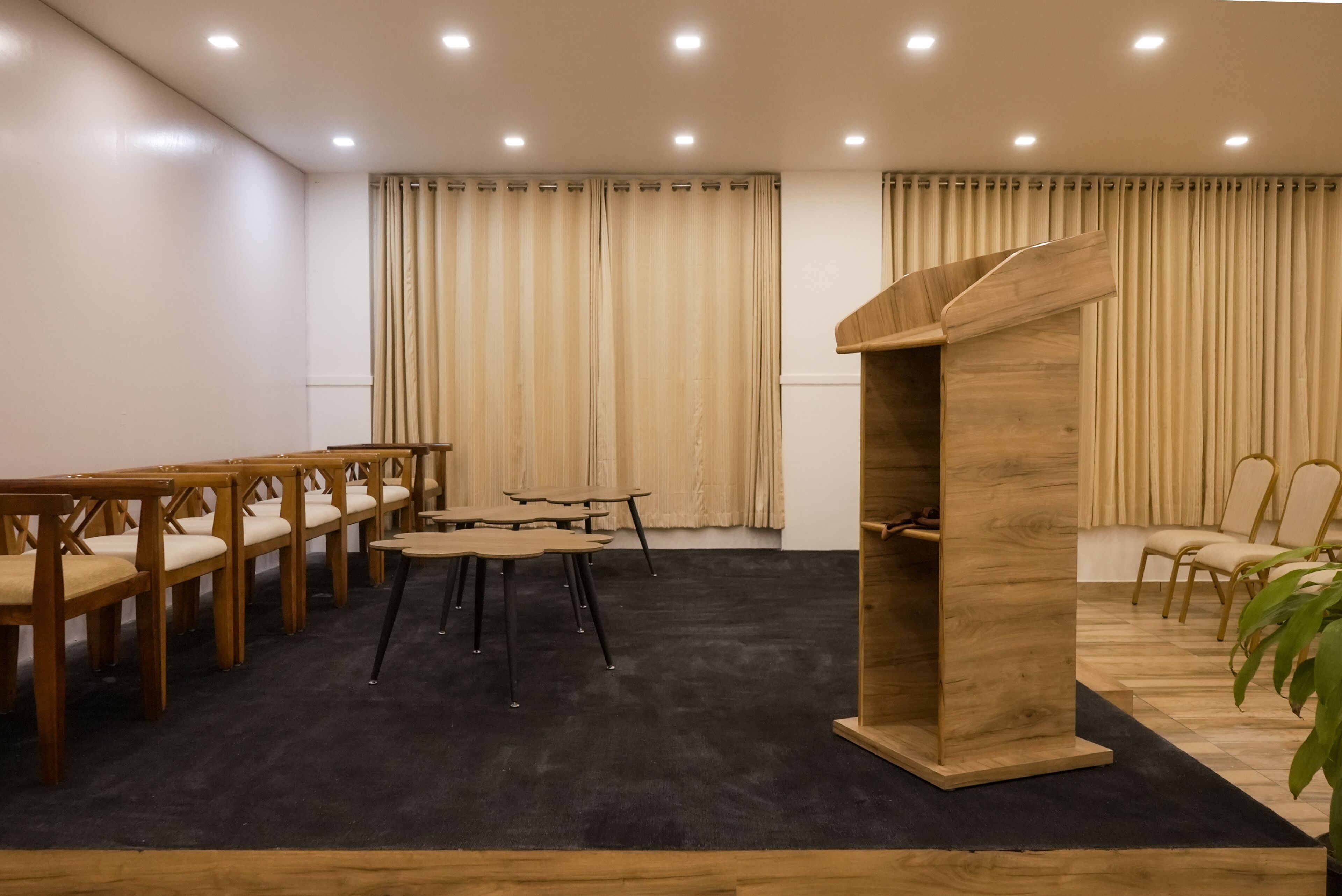 Une salle de conférence avec des tables et des chaises en bois, un pupitre et des rideaux beiges.