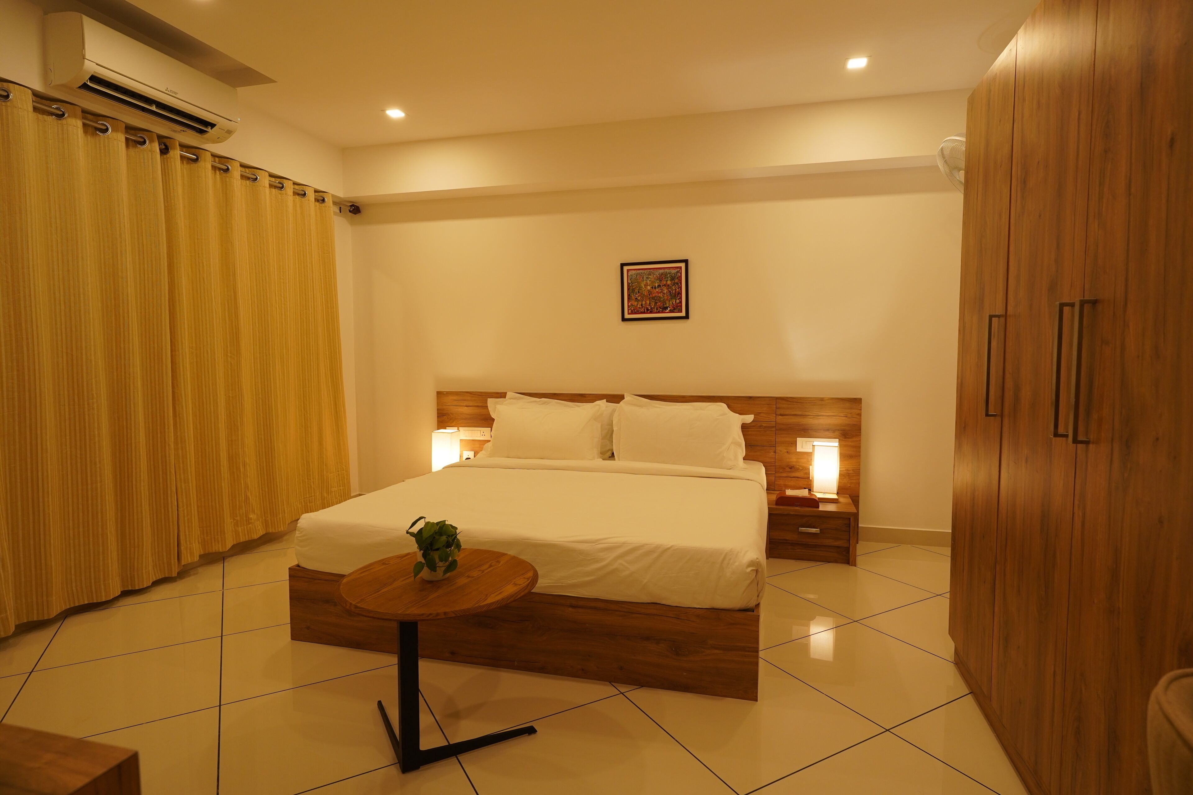 A modern bedroom with a large bed, wooden headboard, bedside tables with lamps, a round wooden table with a plant, and a wardrobe.