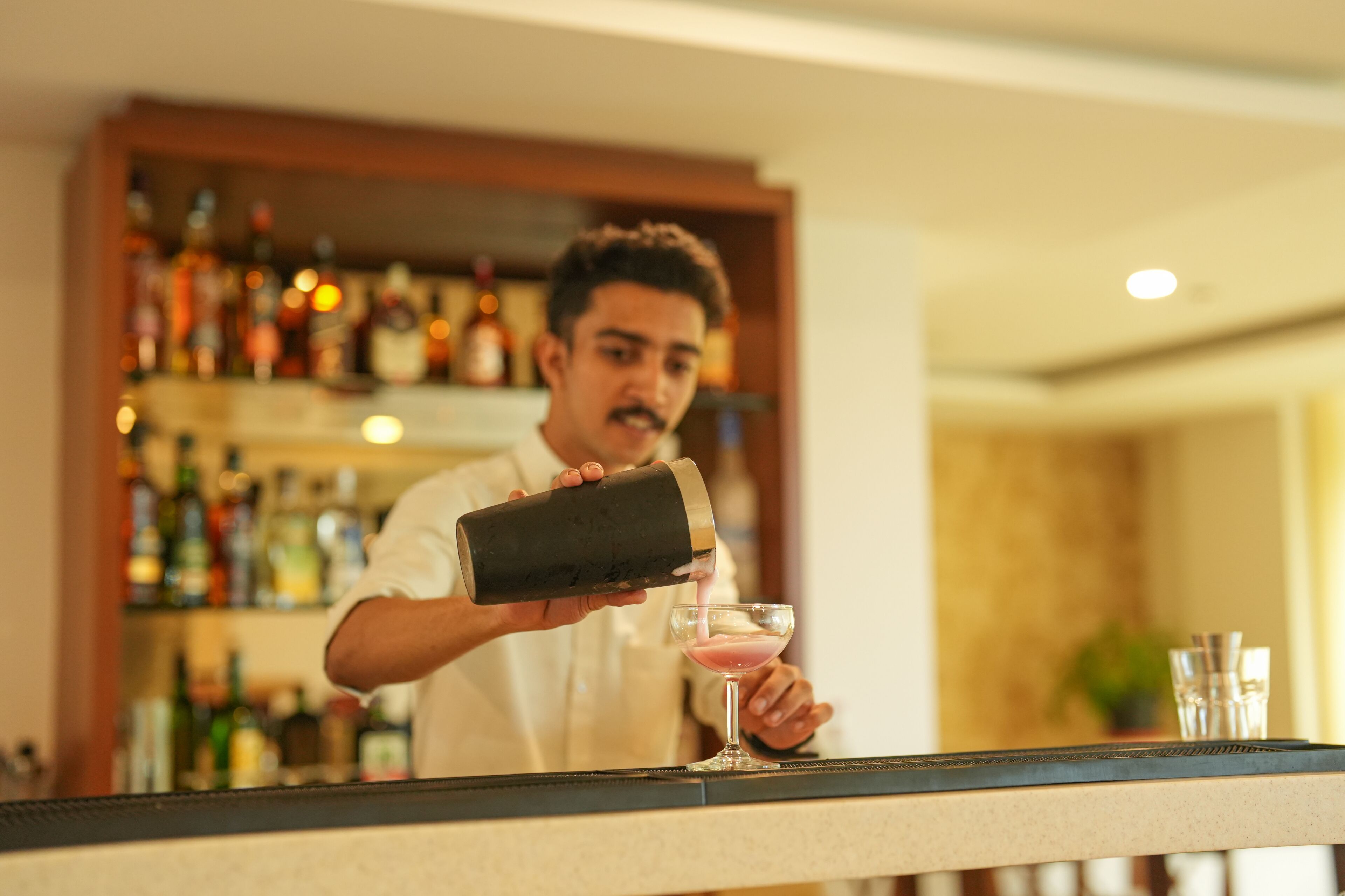 A bartender pouring a drink into a martini glass at a bar with shelves of bottles in the background.