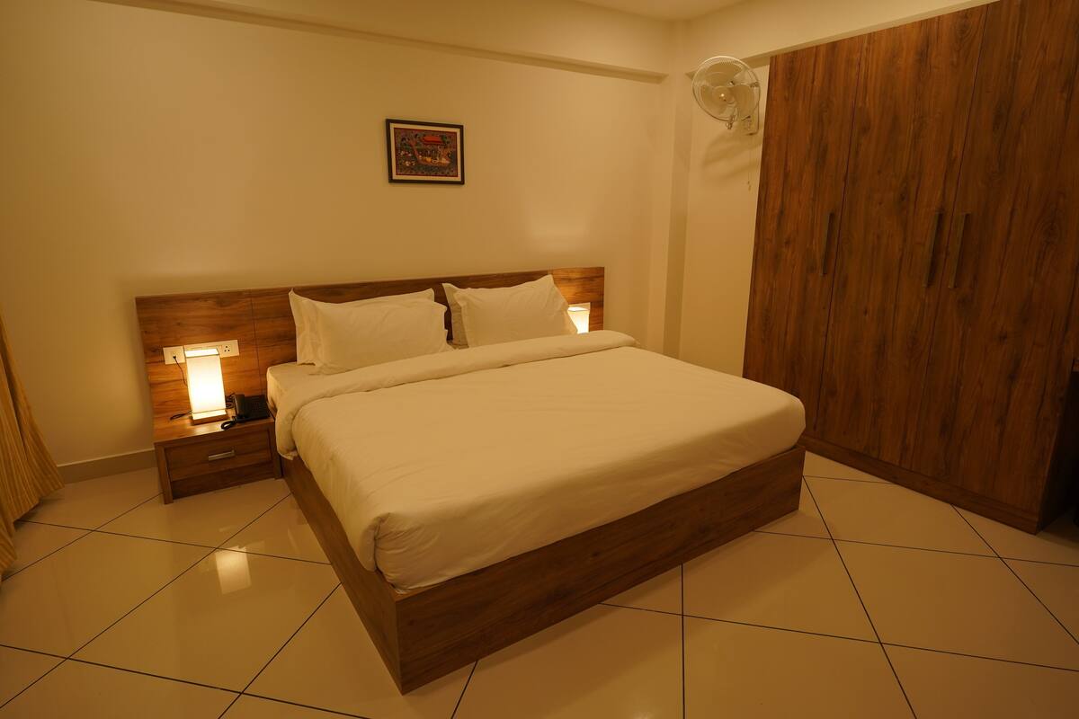 A neatly made bed in a hotel room with a wooden headboard, a nightstand with a lamp, and a wardrobe.