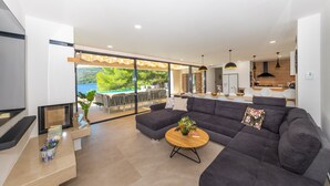 A modern living room with a large dark-colored sectional sofa, a round wooden coffee table, and a TV mounted on the wall. There is a large window providing a view of a pool and greenery outside. - Beachfront Villa Brulupes (Ston)