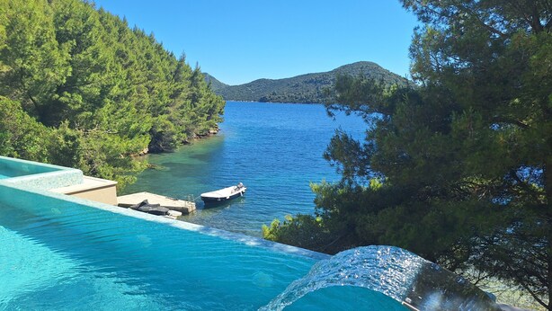 A clear blue pool with a waterfall feature, surrounded by lush greenery and a view of the sea. - Beachfront Villa Brulupes (Ston)