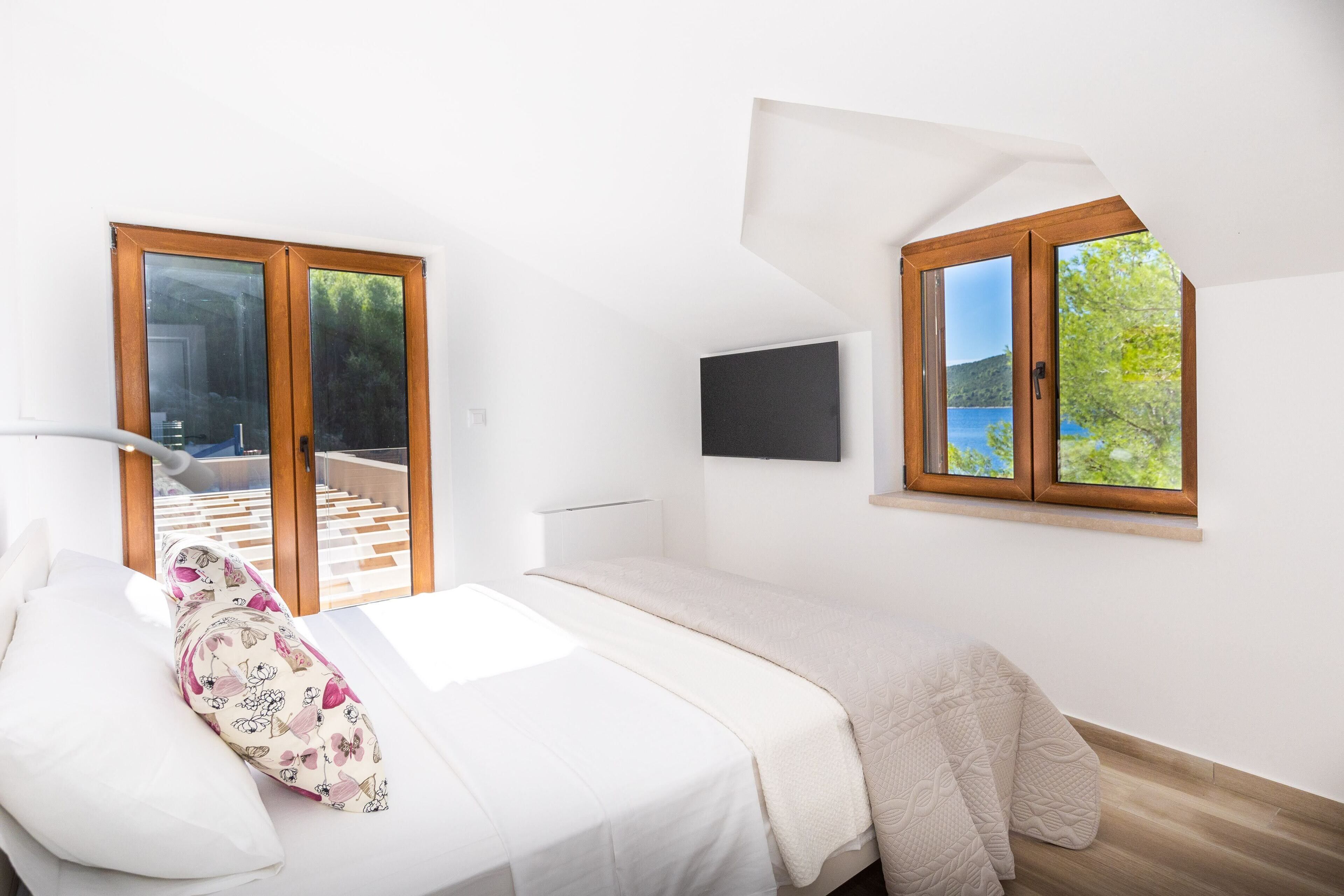 A bedroom with a bed, a window with a view of greenery, and a door leading to an outdoor area.