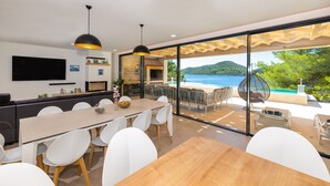 A modern dining area with a large table and chairs, a TV mounted on the wall, and a view of a beach and mountains through large glass doors. - Beachfront Villa Brulupes (Ston)