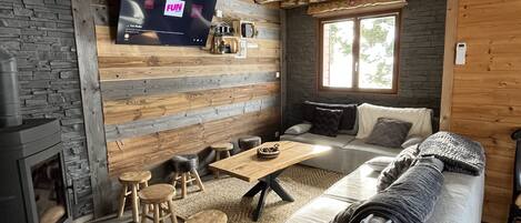 Ein gemütliches Wohnzimmer mit Steinwand, Holzbalken, Kamin, Sofa, Couchtisch und Wandfernseher.