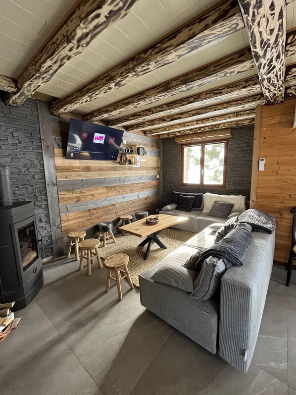 Una sala de estar acogedora con pared de piedra, vigas de madera, chimenea, sofá, mesa de centro y televisor montado en la pared.