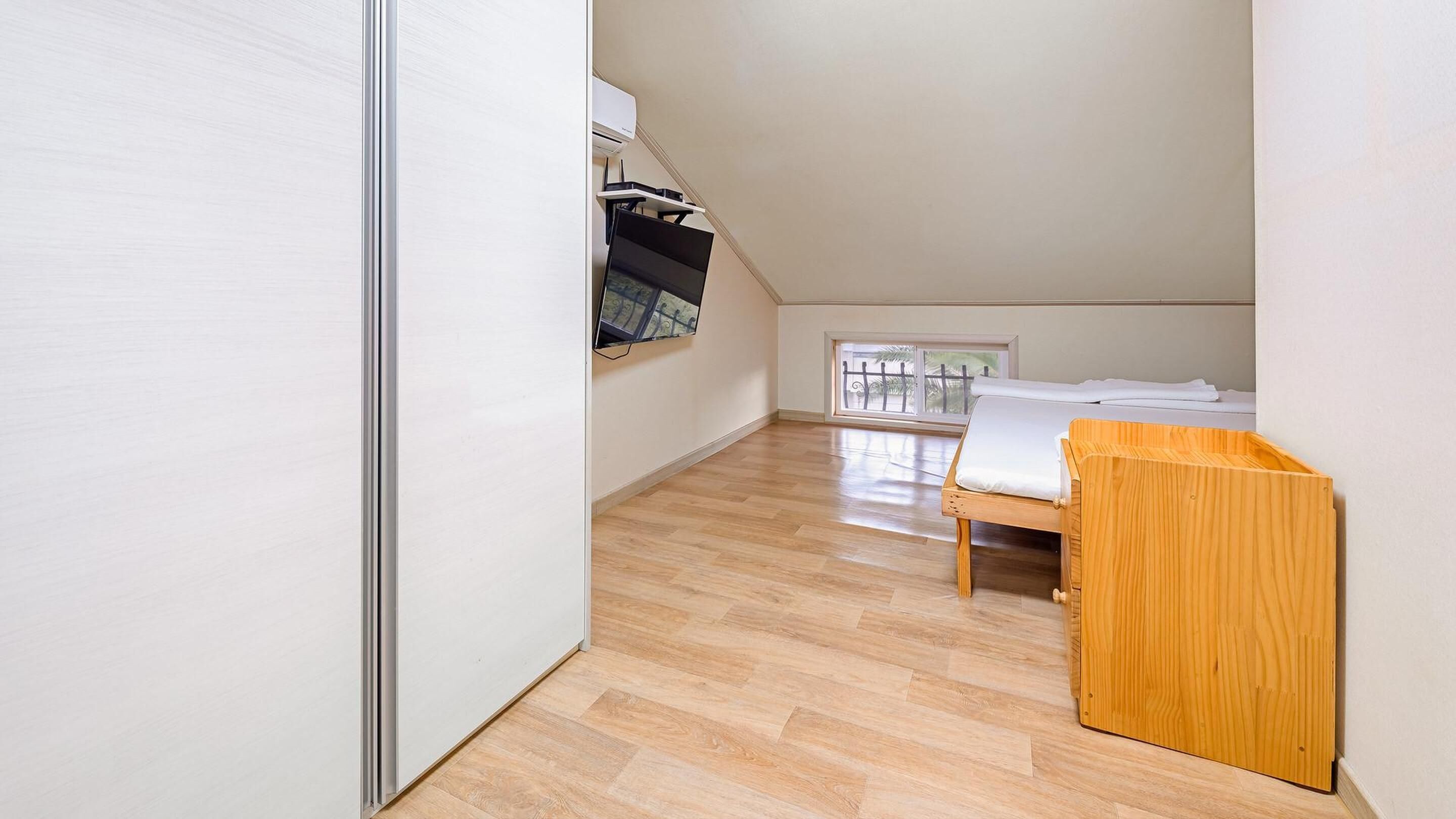 Een kleine, lichte kamer met een houten vloer, een bed, een raam met uitzicht en een kledingkast met daarin hangende kleding.