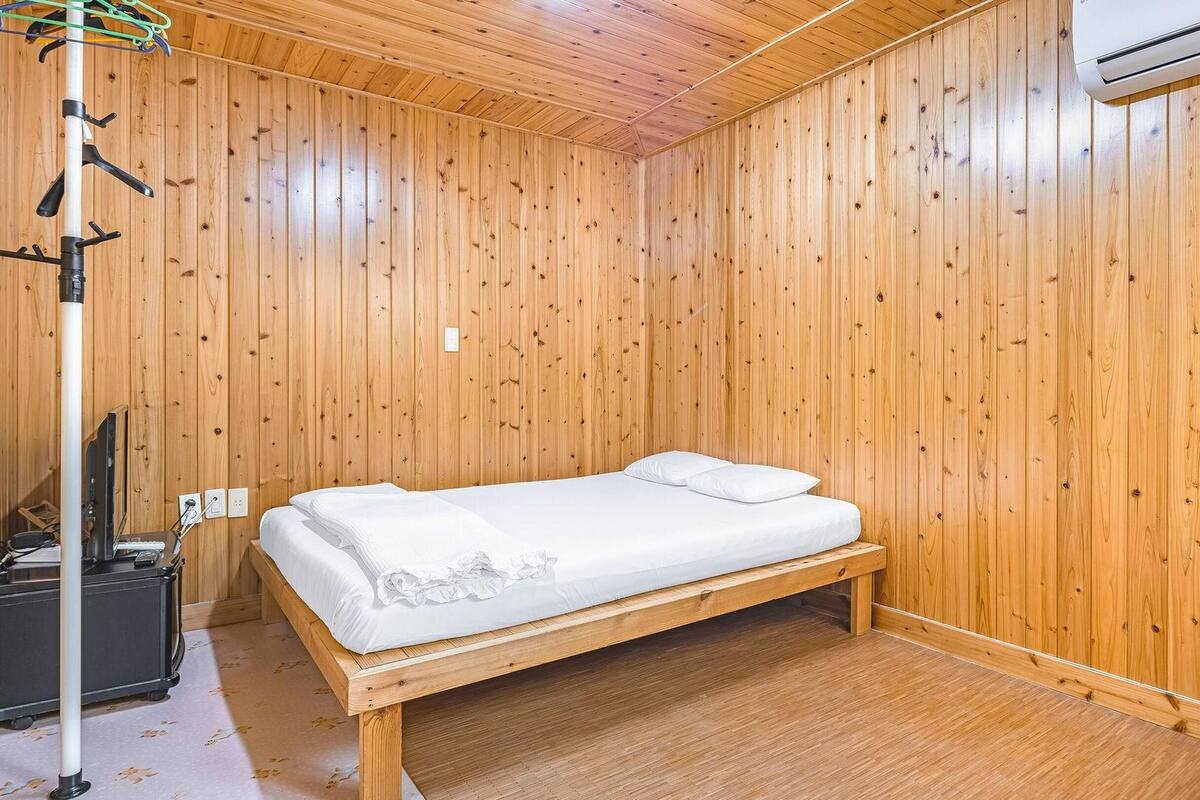 A wooden-paneled room with a single bed, a desk with a computer, and an air conditioning unit.