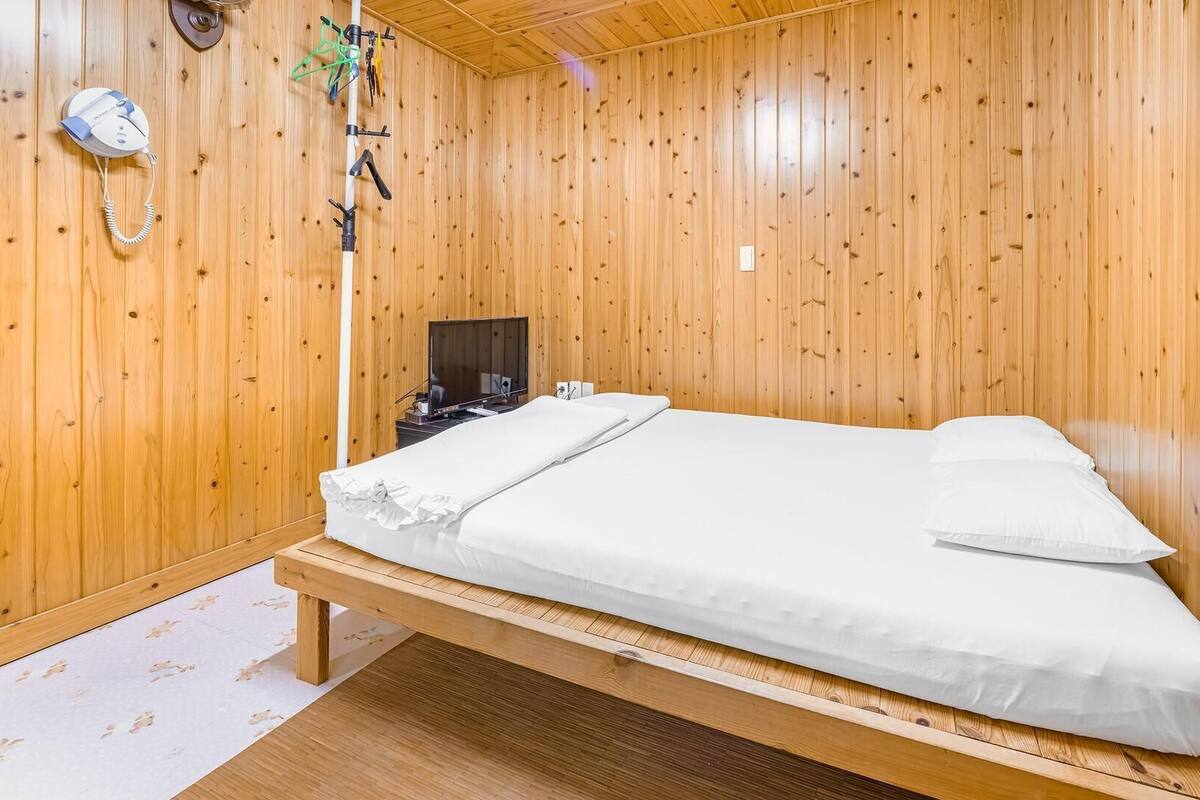 A single-bed room with wooden walls, a bedside table, a television, and a telephone.
