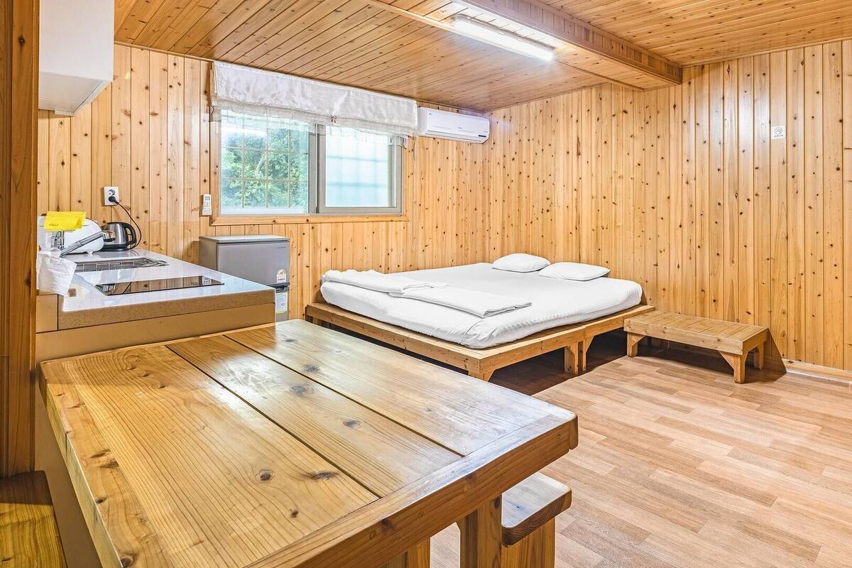A wooden room with a bed, a small table, a bench, and a window.
