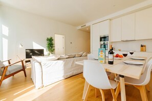 A modern living room with a sofa, a dining table with chairs, a TV, and a kitchen area with cabinets and a plant.