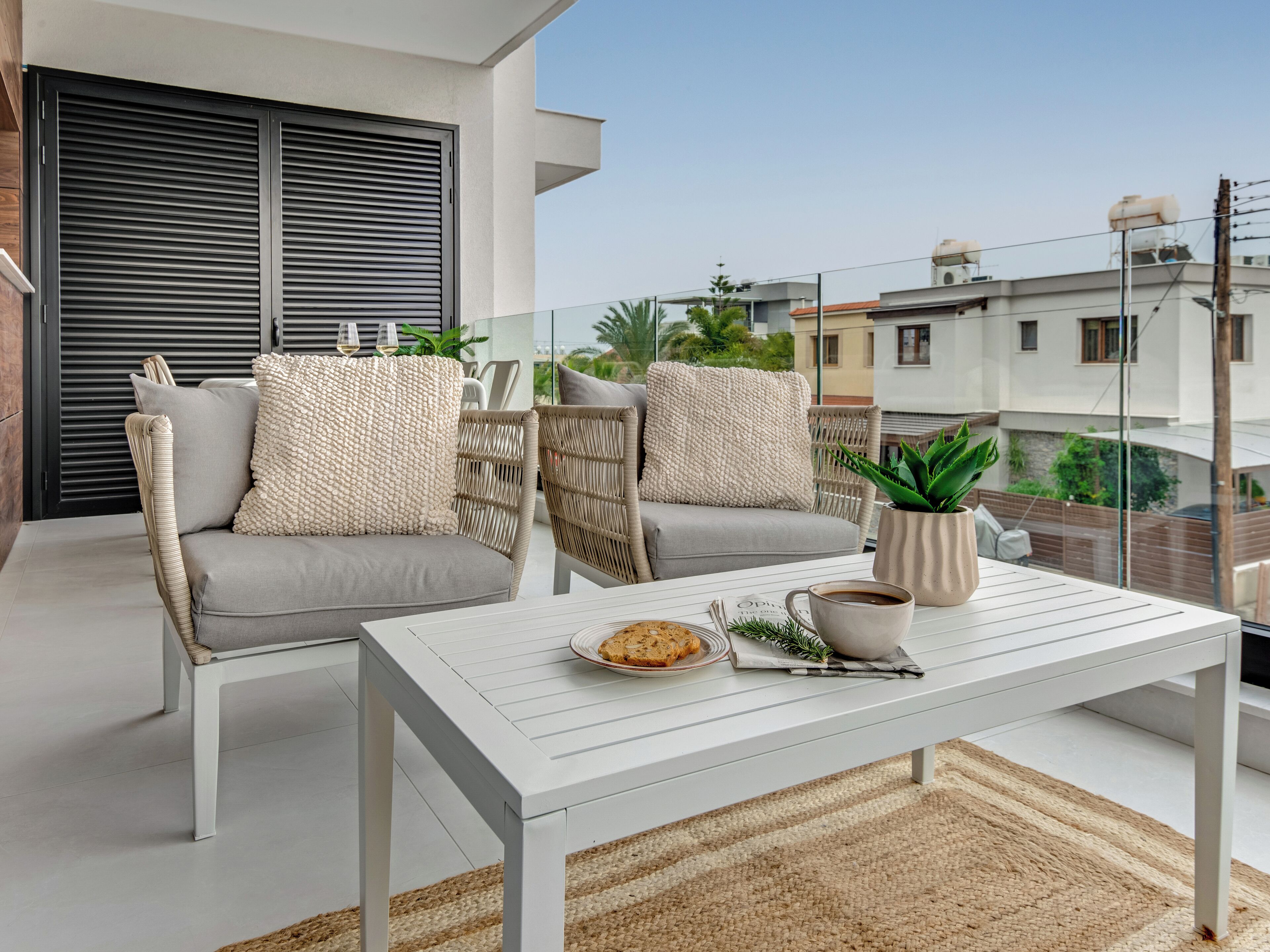 A balcony with a white table, two wicker sofas with cushions, a potted plant, and a view of neighboring buildings.