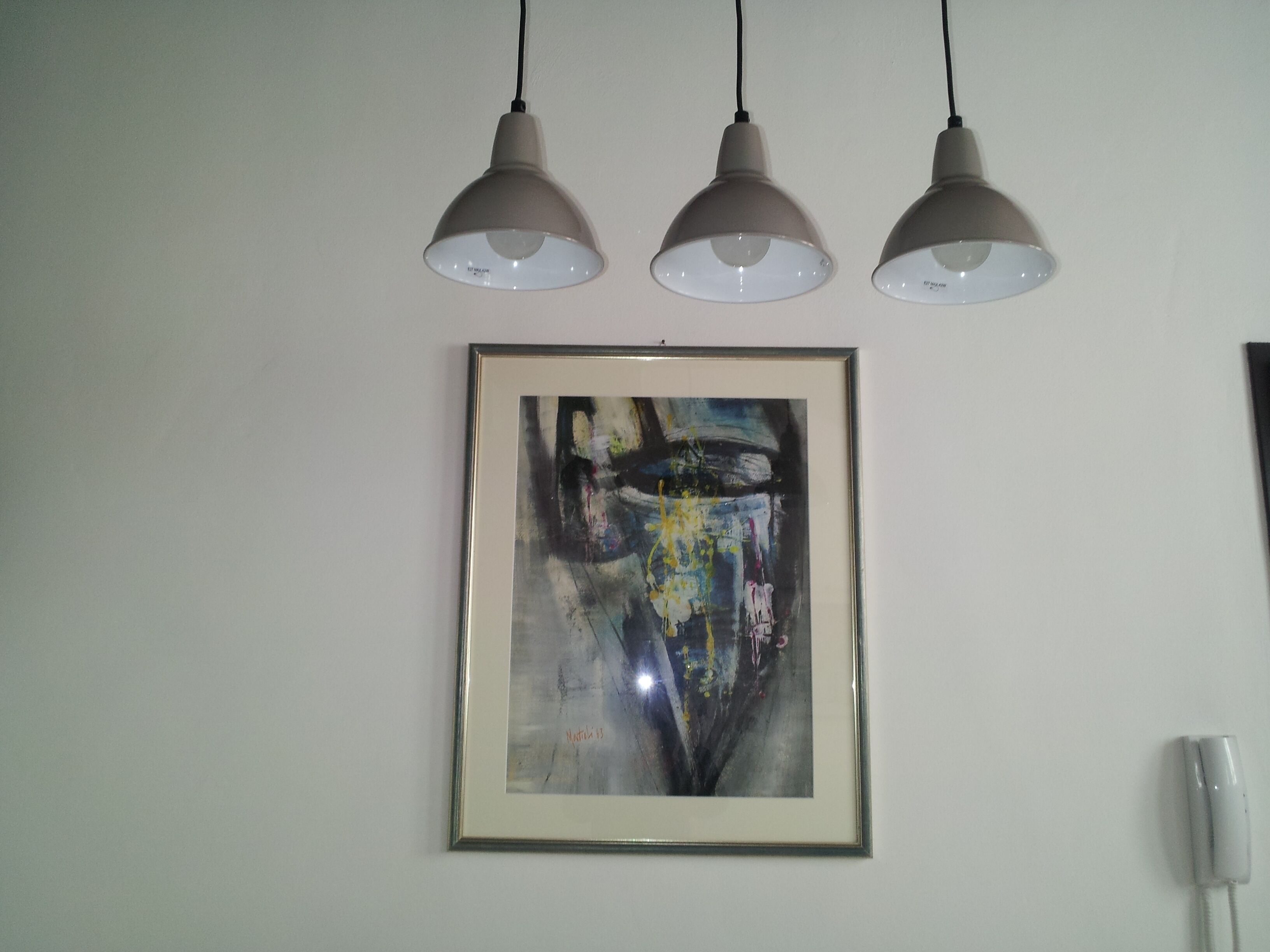 Une peinture abstraite encadrée est accrochée à un mur blanc, et trois suspensions lumineuses sont disposées au-dessus.
