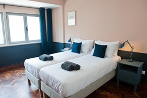 Una cama doble con sábanas blancas y almohadas azul oscuro, dos mesitas de noche con lámparas, una ventana con cortinas y un suelo de madera con patrón de espiga.